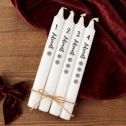 Vier Adventskerzen in Weiß mit Beschriftung und Schneeflocken, dekorativ zusammengebunden auf Holztisch mit rotem Stoff.