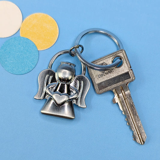 Schlüsselanhänger in Engel-Form mit Herz in der Hand, silberfarben, neben Hausschlüssel auf blauem Hintergrund