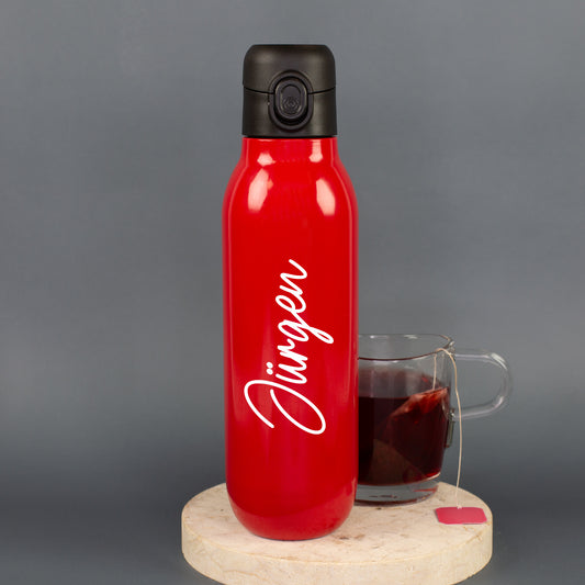 Personalisierte rote Edelstahl-Trinkflasche mit weißem Namen „Jürgen“ neben einer Tasse Tee als Geschenkidee