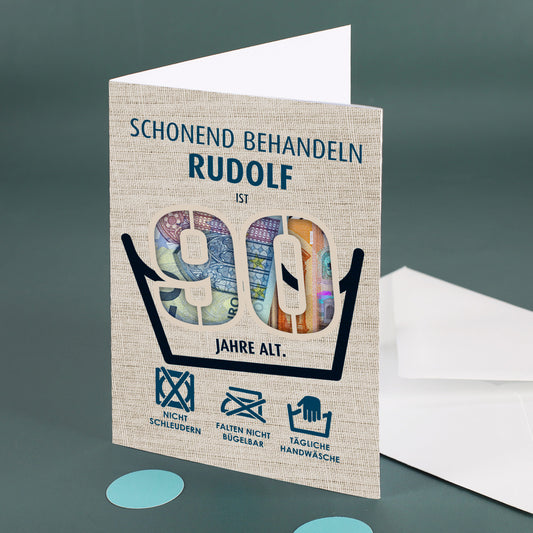 Aufgestellte Klappkarte mit dem Namen Rudolf und der Zahl 90, hinterlegt mit bunten Geldscheinen, daneben ein weißer Umschlag auf grünem Untergrund.