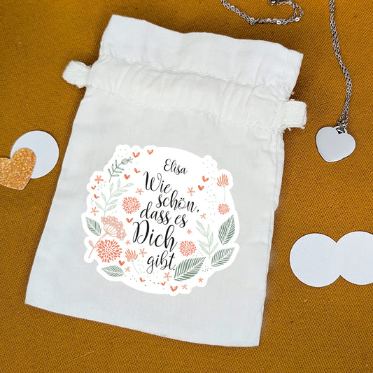 Stoffbeutel mit floraler Botschaft „Wie schön, dass es dich gibt“ – personalisiert mit Name – auf Tisch mit Kette und Deko