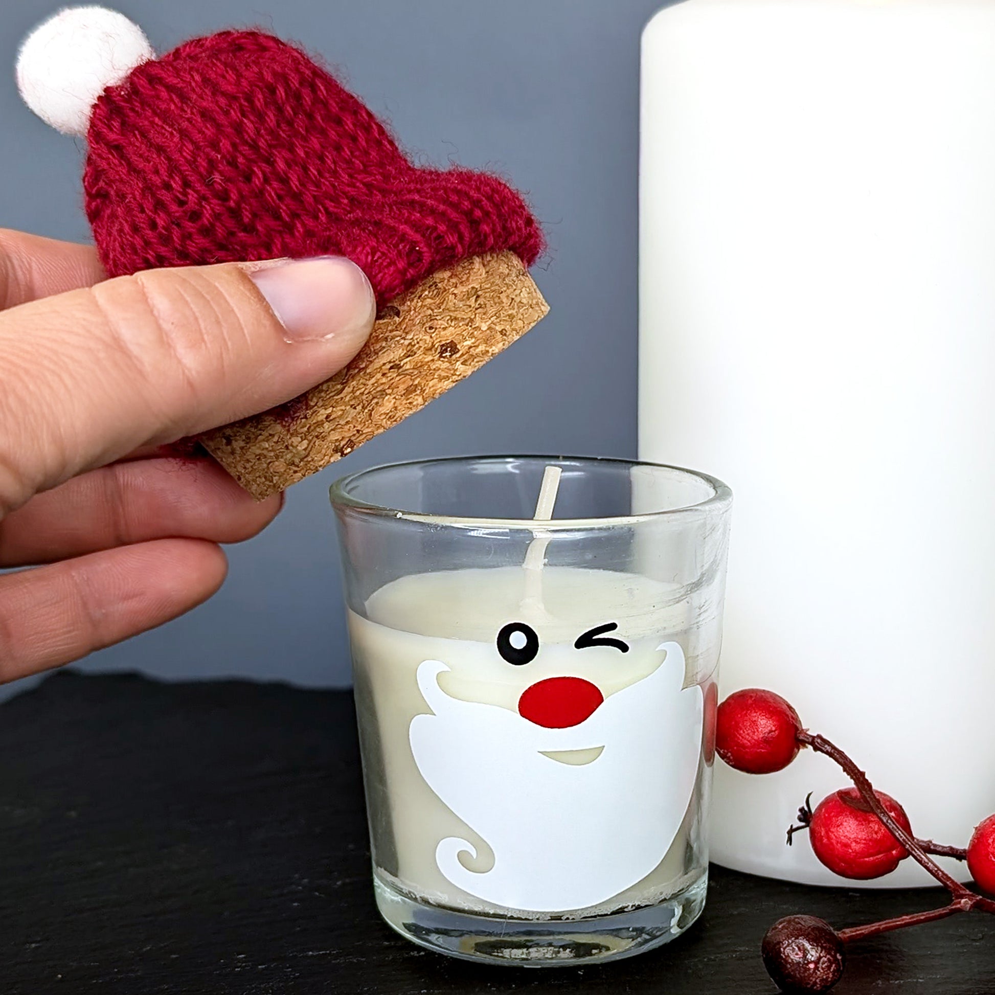 Hand hebt rote Pudelmütze mit Korkverschluss von der Kerze im Glas mit Weihnachtsgesicht – liebevolle Detailaufnahme.