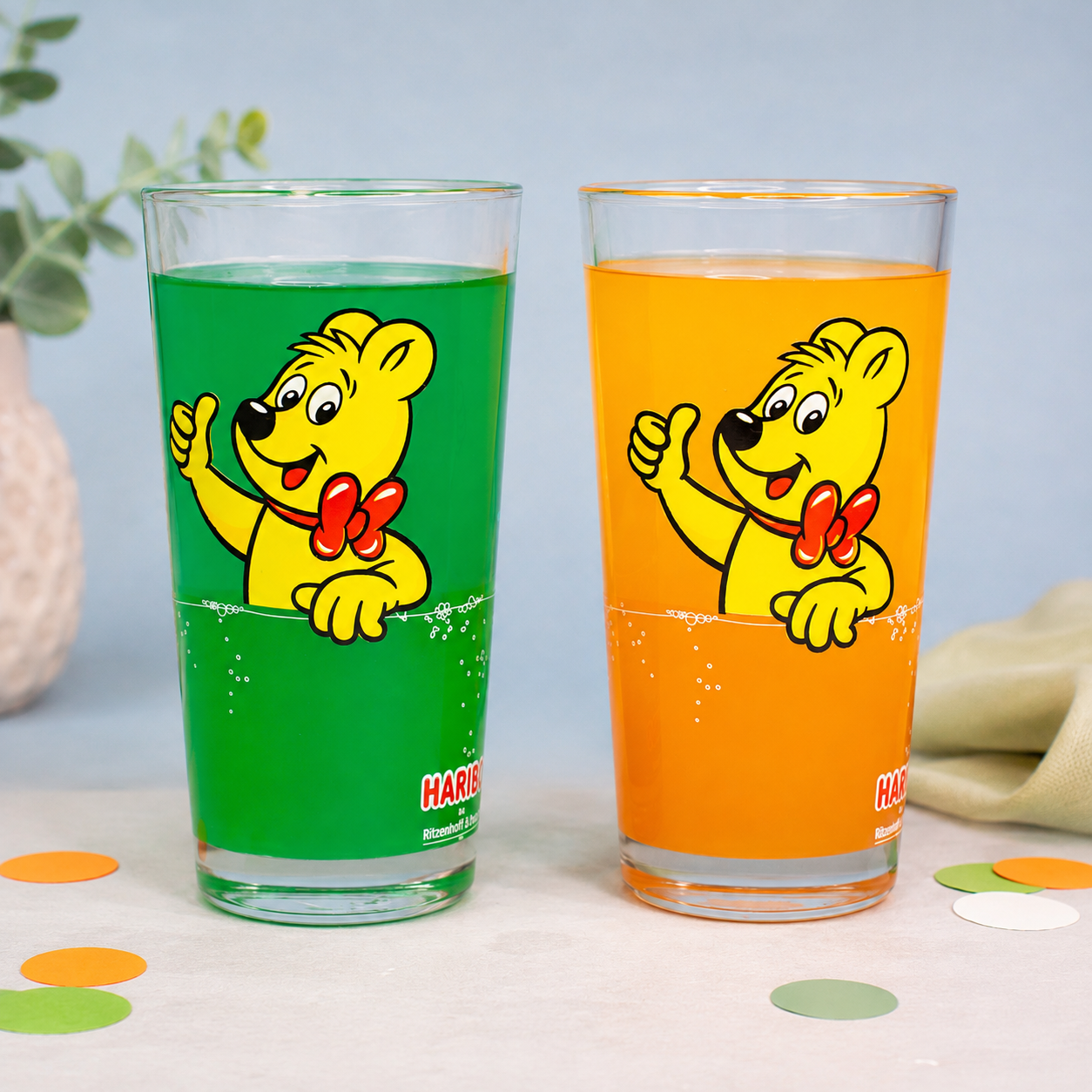 Zwei HARIBO Trinkgläser ohne Personalisierung mit Goldbär Motiv, grünem und orangem Getränk, dekorativ auf hellem Hintergrund arrangiert