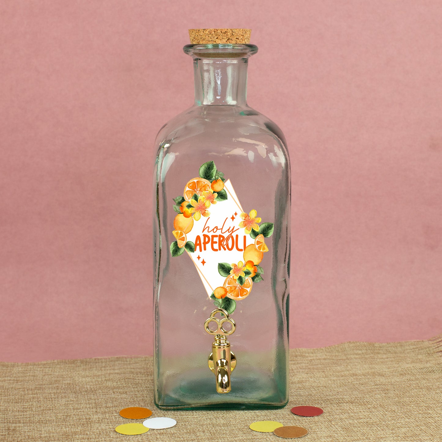 Glas Getränkespender mit Zapfhahn und sommerlichem Orangen-Motiv auf Stoffunterlage vor rosa Hintergrund