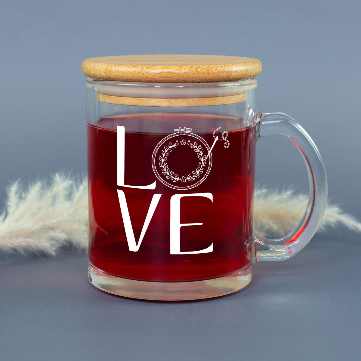 Glastasse mit rotem Tee, LOVE Stickmotiv und aufgesetztem Bambusdeckel vor grauem Hintergrund