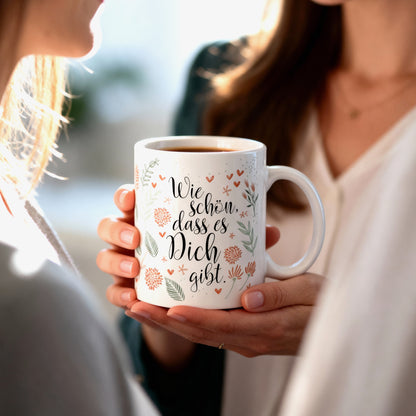 Frau hält Tasse mit Aufdruck „Wie schön, dass es Dich gibt“ in den Händen und schenkt sie einer anderen Person.