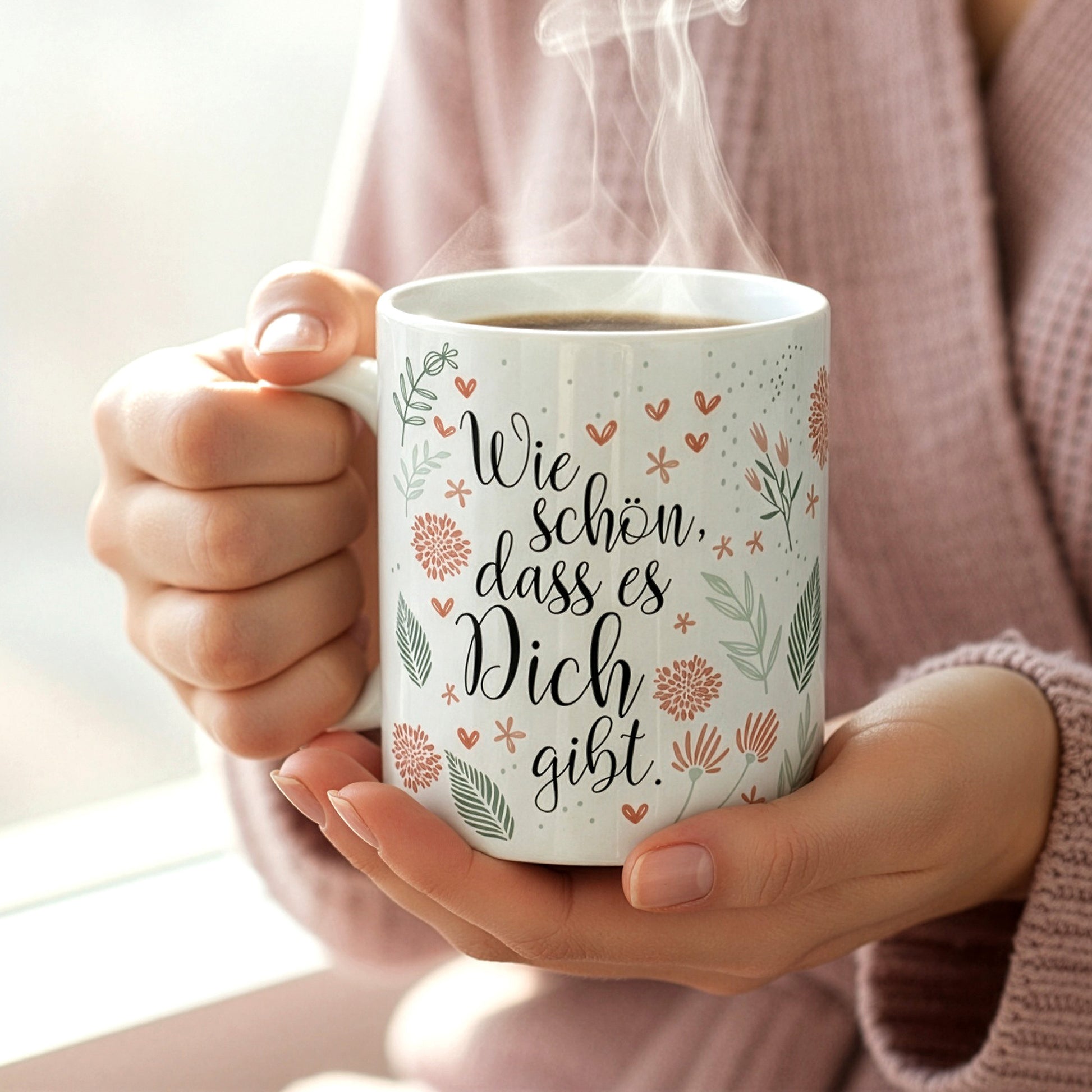 Person hält dampfende Tasse mit Aufdruck „Wie schön, dass es Dich gibt“ in beiden Händen, im Hintergrund weiches Tageslicht.