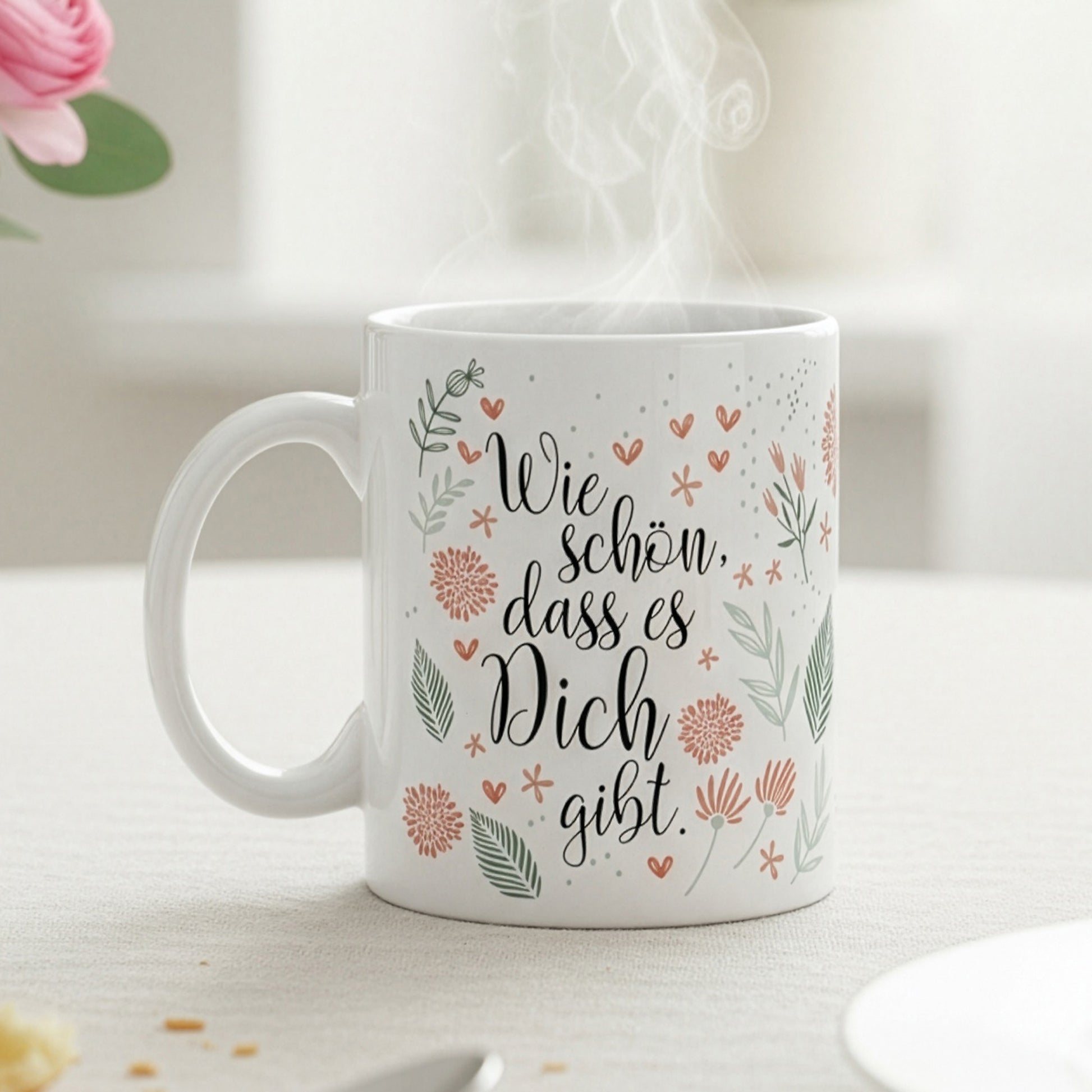 Tasse mit Blumenmotiv und Schriftzug „Wie schön, dass es Dich gibt“ auf Frühstückstisch mit rosa Blume und Croissant.