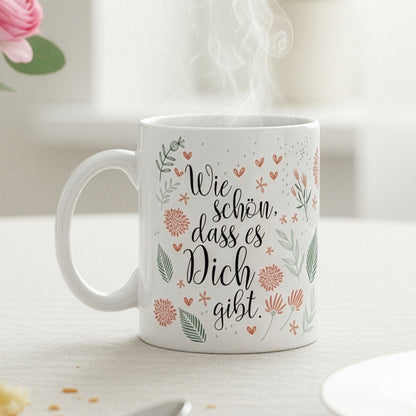 Tasse mit Blumenmotiv und Schriftzug „Wie schön, dass es Dich gibt“ auf Frühstückstisch mit rosa Blume und Croissant.