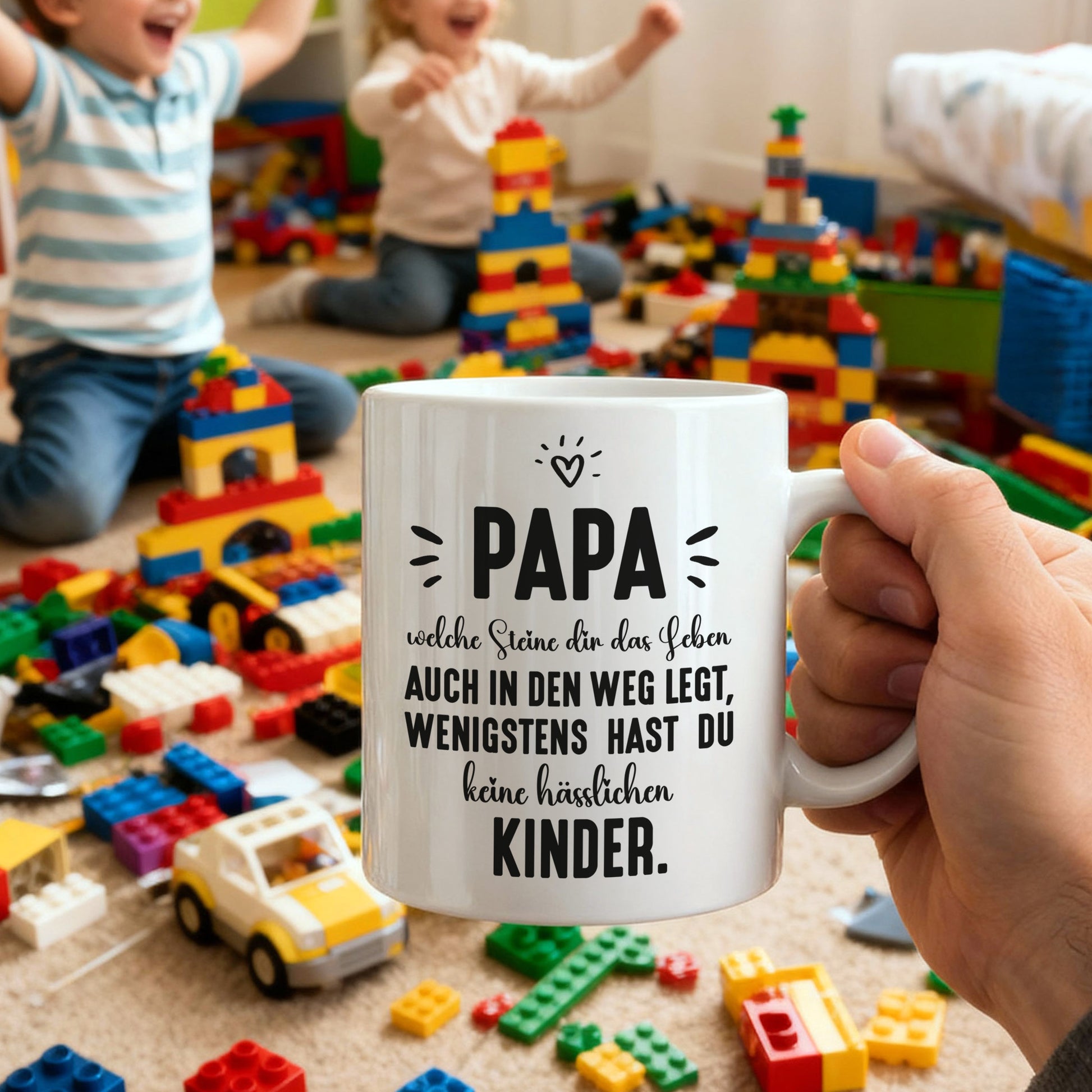 Papa-Tasse mit humorvollem Spruch, gehalten in der Hand, im Hintergrund Kinder mit bunten Bausteinen