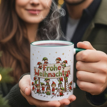 Person hält eine dampfende Weihnachtstasse mit werkelnden Wichteln am „Frohe Weihnachten“-Schild; Paar im Hintergrund leicht unscharf.