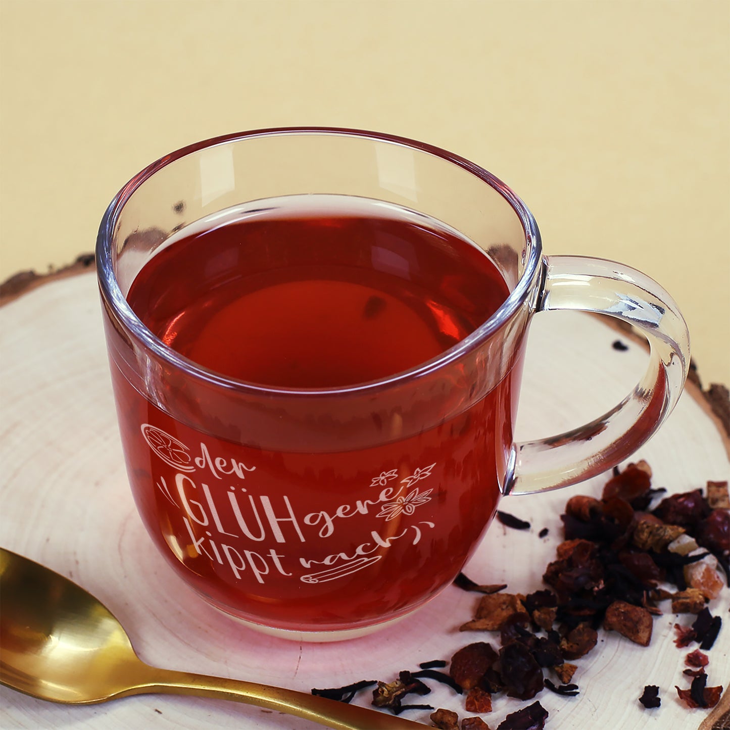 Leonardo Glühweintasse „Der GLÜHgere kippt nach“ auf Holzscheibe, befüllt mit Glühwein, mit goldfarbenem Löffel daneben