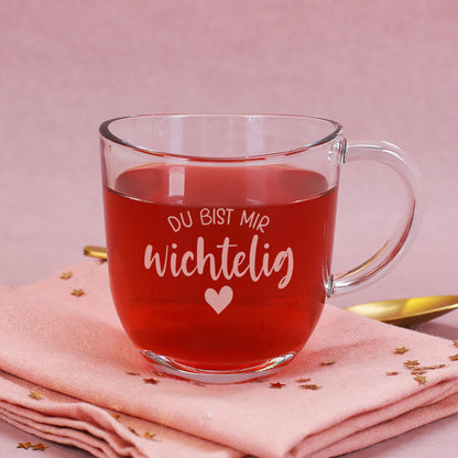 Teeglas mit der Gravur „Du bist mir wichtelig“ auf rosa Untergrund, gefüllt mit rotem Tee.