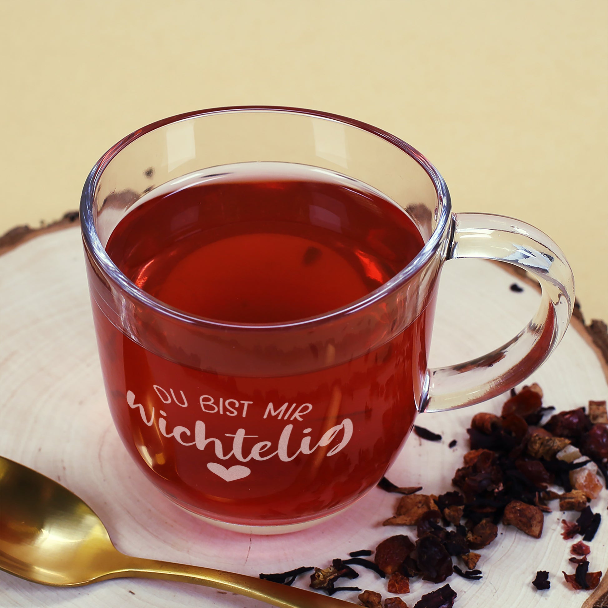 Teeglas mit graviertem Spruch „Du bist mir wichtelig“ auf Holzscheibe, daneben goldener Löffel und Teemischung.