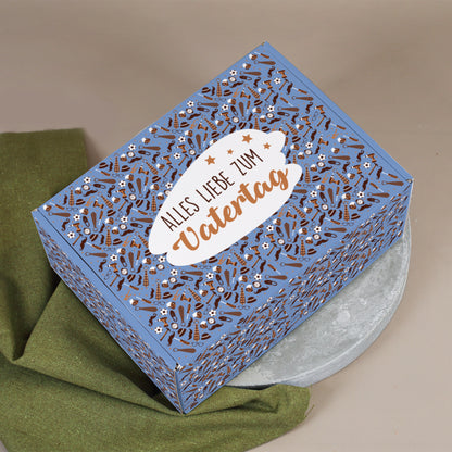 Geschenkbox mit Aufdruck „Alles Liebe zum Vatertag“, dekorativ fotografiert auf grünem Tuch und hellem Teller