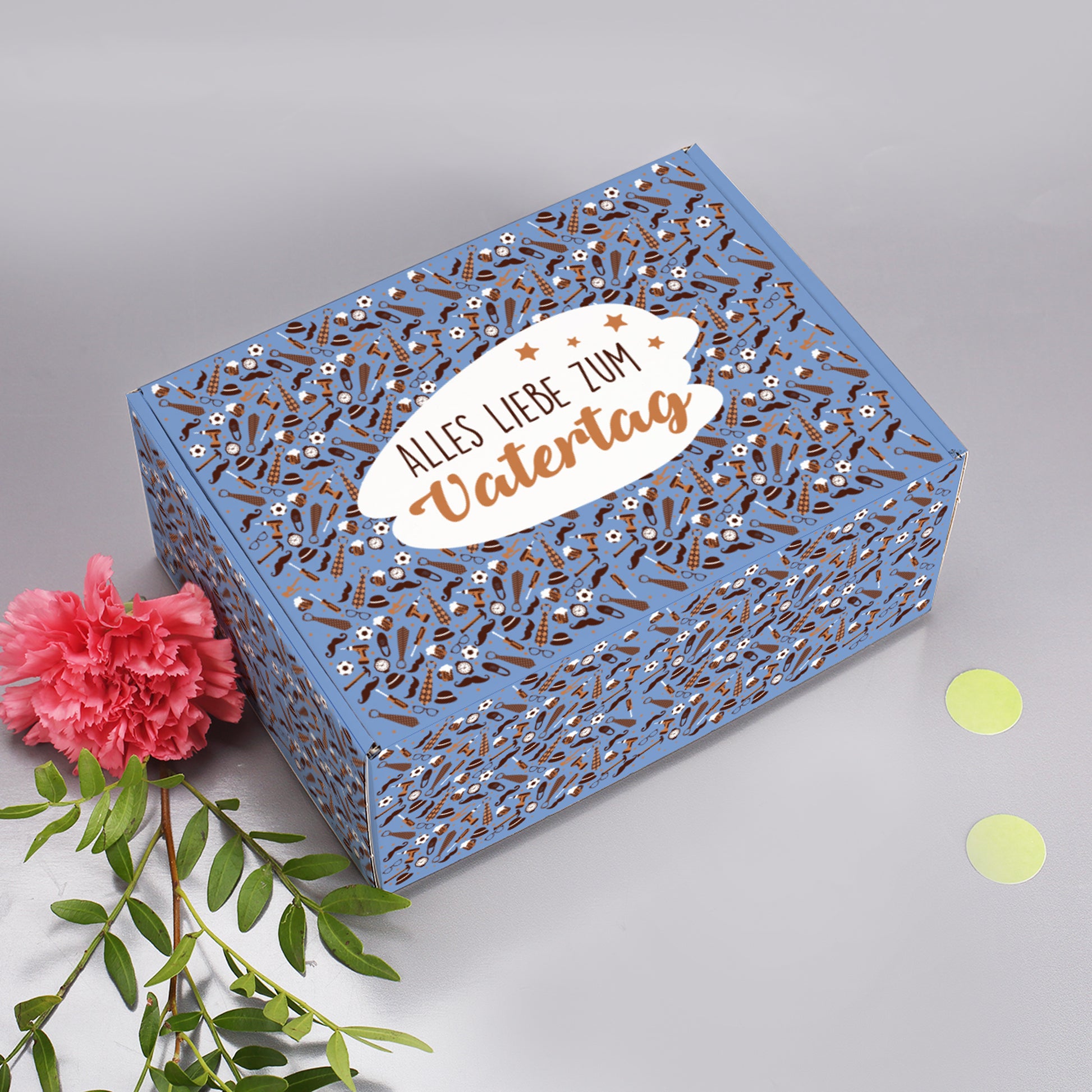 Vatertagsbox mit liebevollem Design, daneben rosa Nelke und Blattzweig zur Dekoration