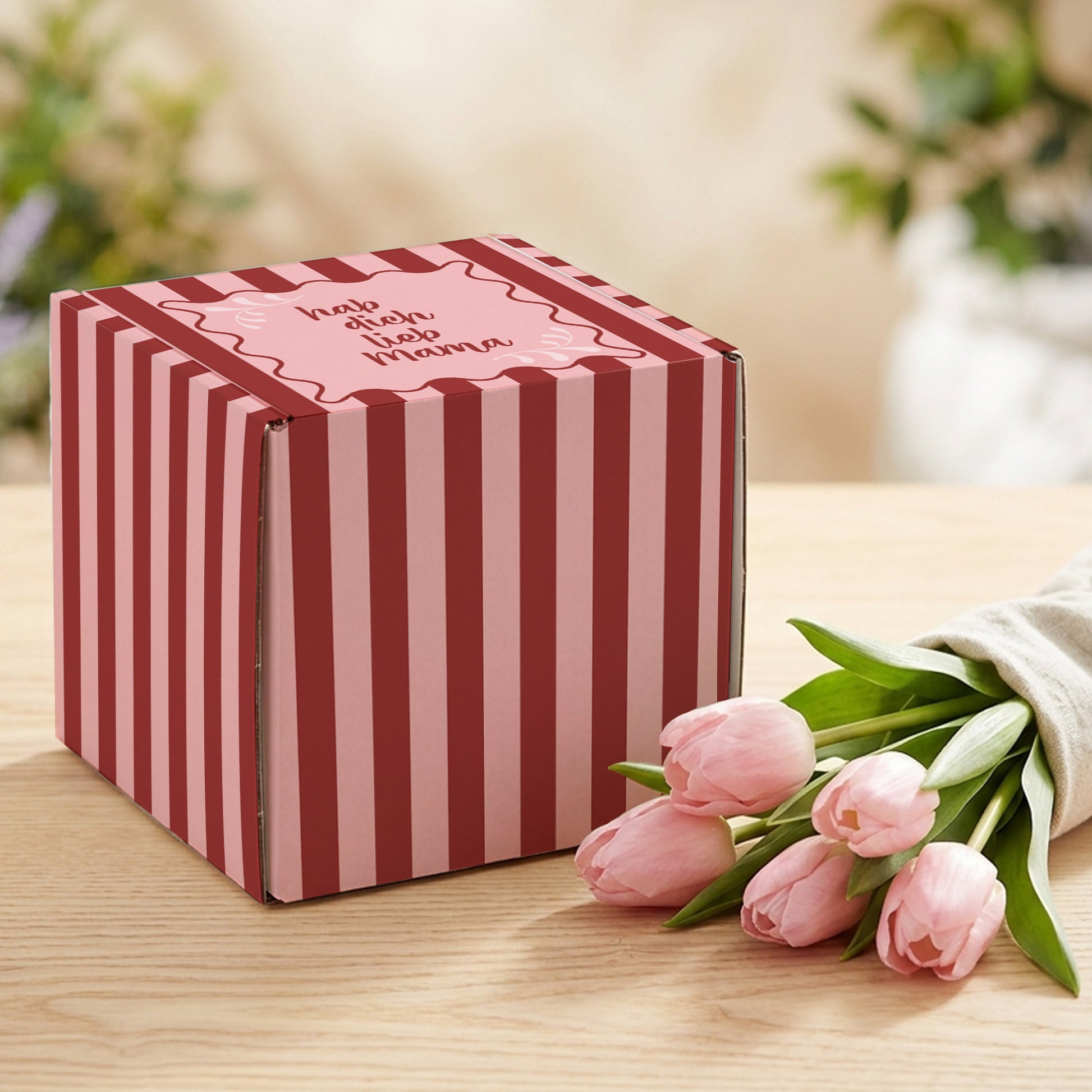 Geschenkbox zum Muttertag mit rosa-rotem Streifenmuster und Spruch „hab dich lieb Mama“ auf hellem Holztisch neben rosa Tulpen