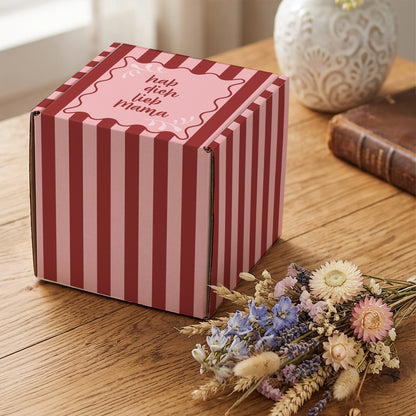 Muttertags-Geschenkverpackung mit Streifenmuster und Aufdruck „hab dich lieb Mama“ auf Holztisch neben Trockenblumen und Deko