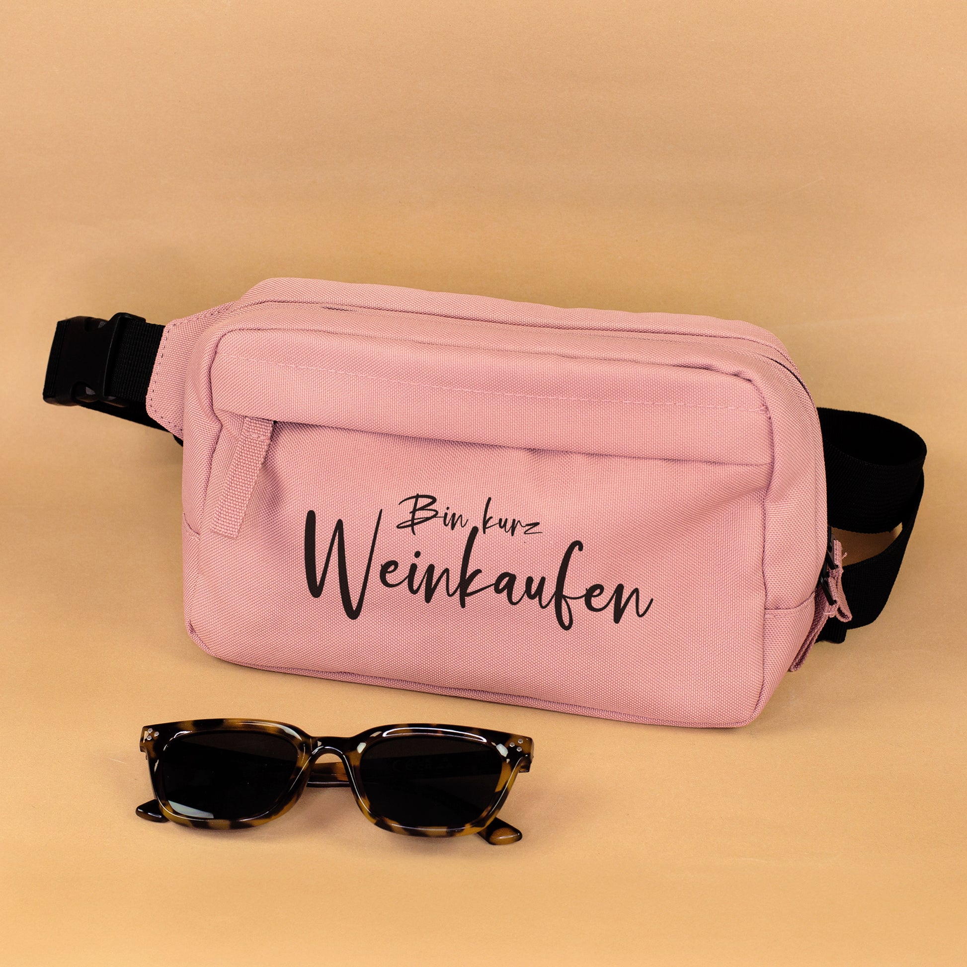 Rosa Bauchtasche mit Spruch Bin kurz Weinkaufen und schwarzem Gurt mit Sonnenbrille als Deko