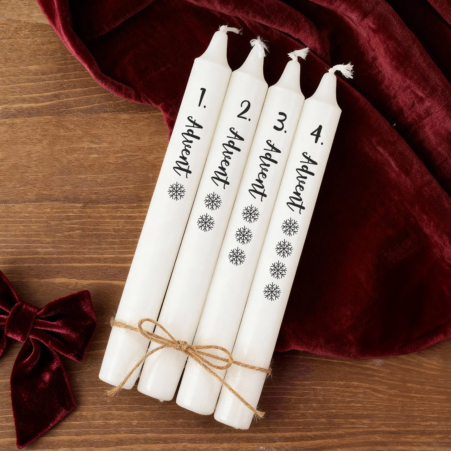 Vier Adventskerzen in Weiß mit Beschriftung und Schneeflocken, dekorativ zusammengebunden auf Holztisch mit rotem Stoff.