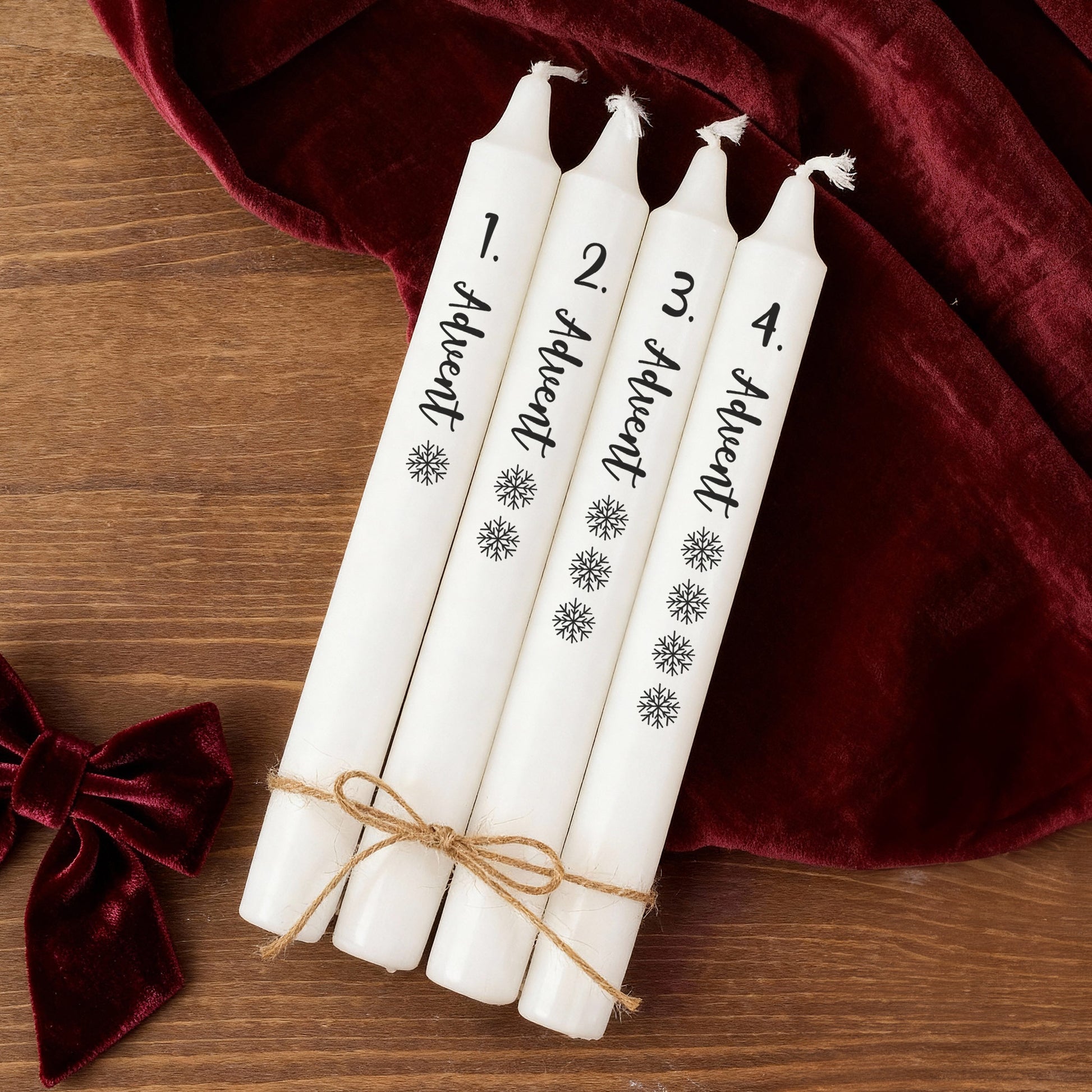 Vier Adventskerzen in Weiß mit Beschriftung und Schneeflocken, dekorativ zusammengebunden auf Holztisch mit rotem Stoff.