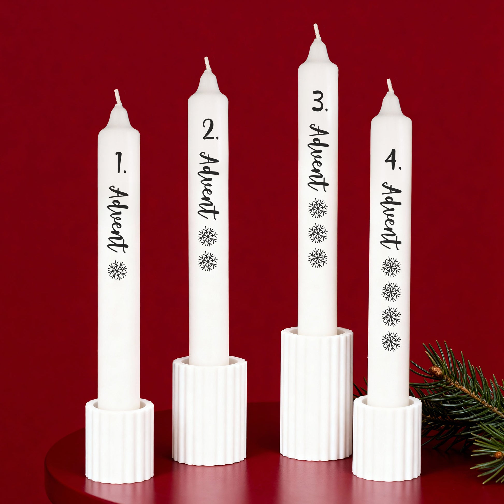 Vier weiße Adventskerzen mit schwarzer Schrift in Kerzenhaltern vor rotem Hintergrund arrangiert.