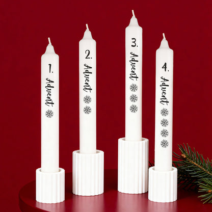 Vier weiße Adventskerzen mit schwarzer Schrift in Kerzenhaltern vor rotem Hintergrund arrangiert.