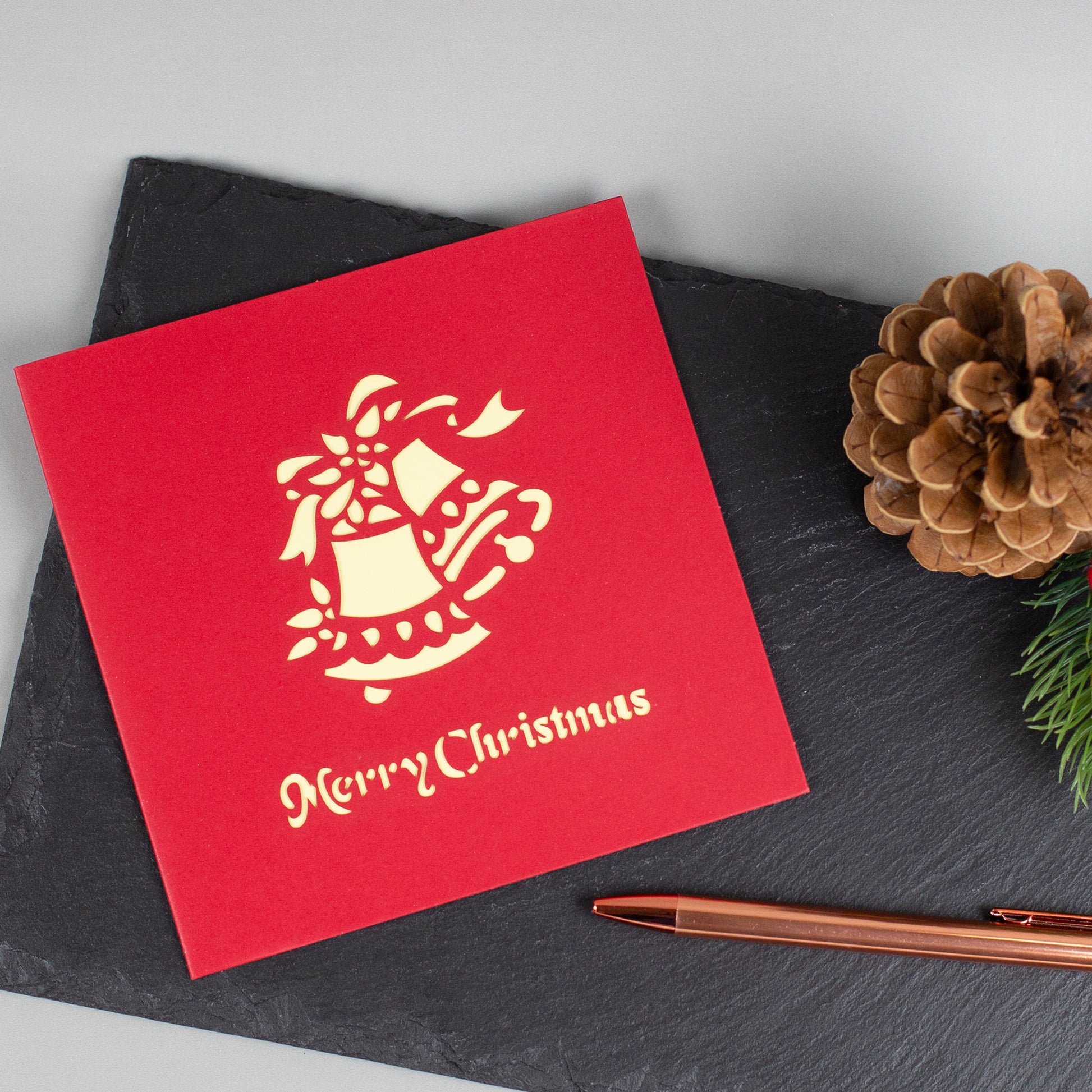 Rote Weihnachtskarte mit goldenem Glockenmotiv und „Merry Christmas“-Schriftzug, dekorativ auf Schieferplatte arrangiert.