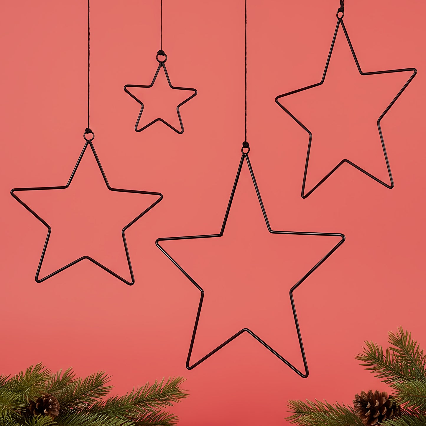 Vier schwarze Metallsterne in verschiedenen Größen hängen dekorativ vor rotem Hintergrund. Im unteren Bereich sind grüne Tannenzweige mit Zapfen zu sehen.
