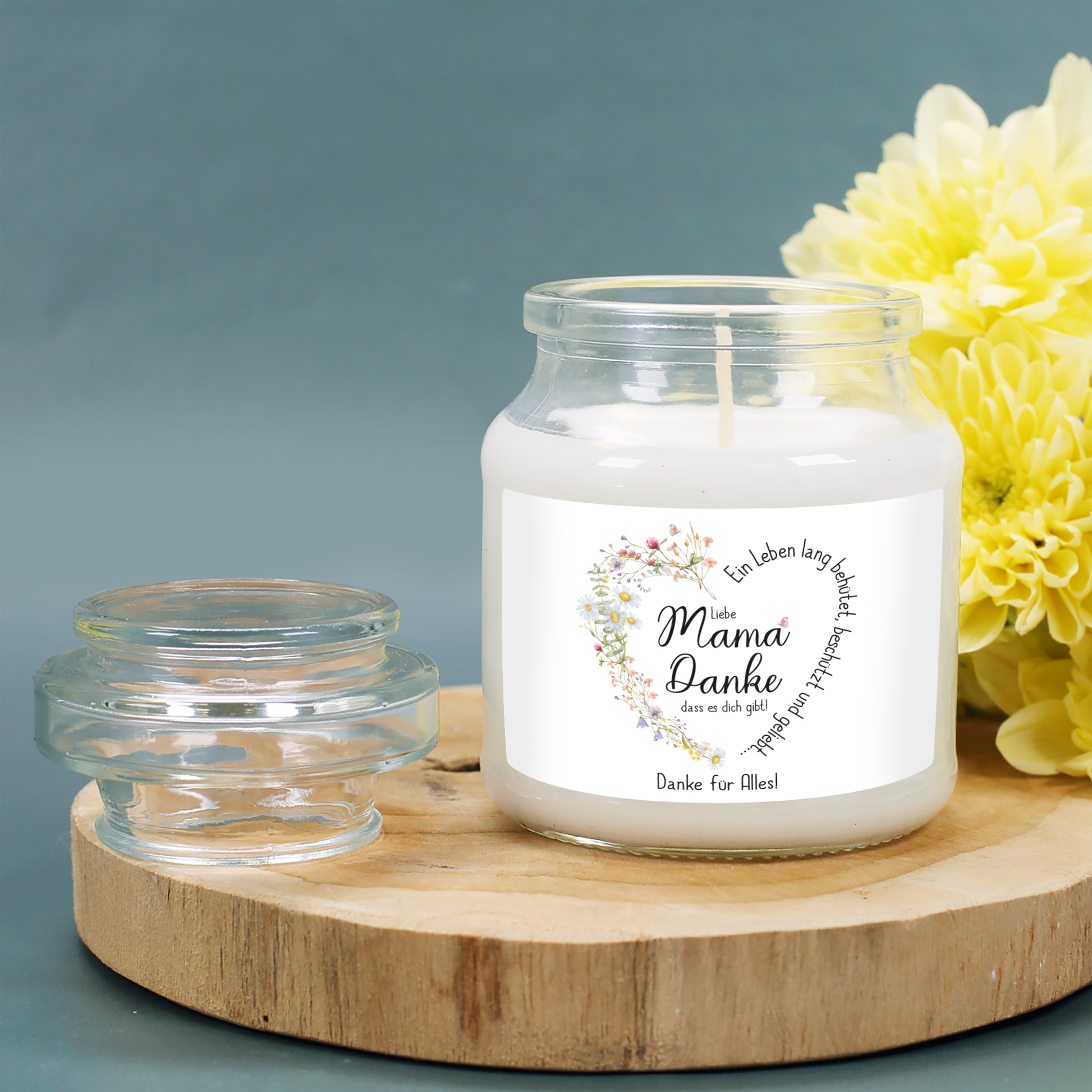 Duftkerze im transparenten Glas mit Deckel, stehend auf einer runden Holzplatte. Die Kerze ist von gelben Blumen umgeben und verbreitet eine warme, gemütliche Atmosphäre. Das Etikett zeigt ein filigranes Herz aus Wiesenblumen, das sich um den Spruch „Ein Leben lang behütet, beschützt und geliebt…“ windet. Die individuelle Personalisierung darunter lautet „Danke für Alles!“. Wunderschönes Muttertags- oder Geburtstagsgeschenk.