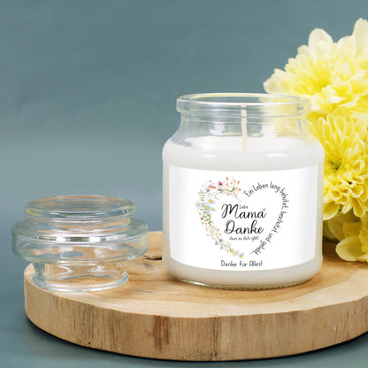 Duftkerze im transparenten Glas mit Deckel, stehend auf einer runden Holzplatte. Die Kerze ist von gelben Blumen umgeben und verbreitet eine warme, gemütliche Atmosphäre. Das Etikett zeigt ein filigranes Herz aus Wiesenblumen, das sich um den Spruch „Ein Leben lang behütet, beschützt und geliebt…“ windet. Die individuelle Personalisierung darunter lautet „Danke für Alles!“. Wunderschönes Muttertags- oder Geburtstagsgeschenk.