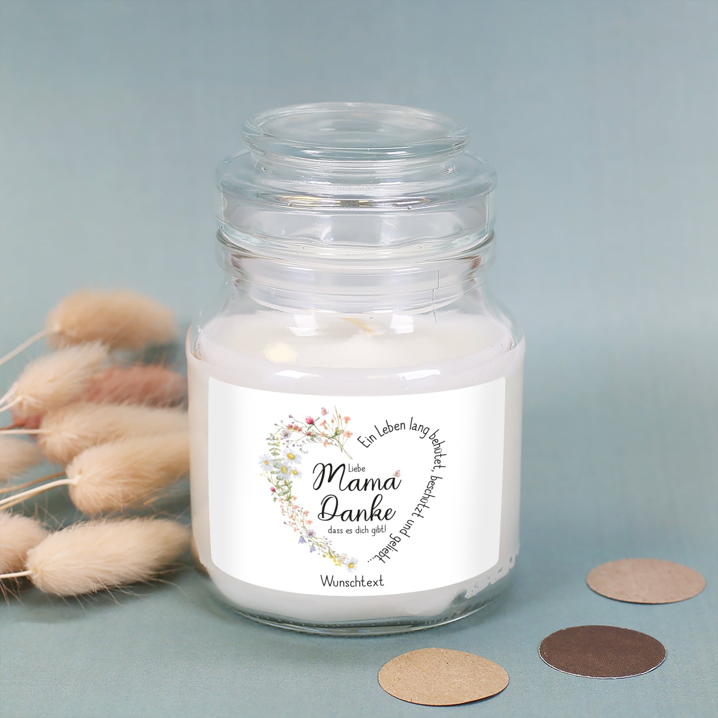 Duftkerze im transparenten Glas mit Deckel, auf einer blauen Stoffunterlage dekoriert mit getrockneten Gräsern und Pappkonfetti in Naturtönen. Das Etikett zeigt ein filigranes Herz aus Wiesenblumen mit dem Spruch „Ein Leben lang behütet, beschützt und geliebt…“ in geschwungener Schrift. In der Mitte steht „Liebe Mama, Danke, dass es dich gibt!“. Unten befindet sich eine Platzhalter-Textzeile mit „Wunschtext“, die für eine individuelle Botschaft personalisiert werden kann.