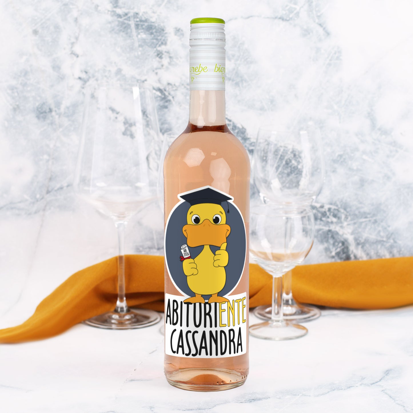 Personalisierte Weinflasche mit Roséwein, Etikett „Abituriente Cassandra“ und Ente mit Doktorhut und Diplom, dekoriert mit Gläsern und orangem Tuch