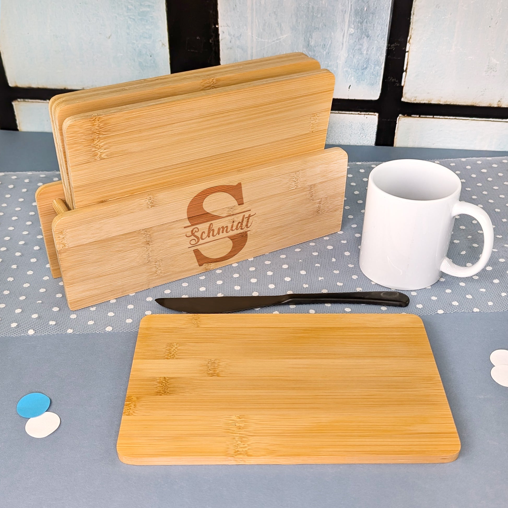 Brettchenständer mit fünf Bambusbrettchen, graviert mit Initiale „S“ und dem Namen „Schmidt“, daneben Messer und weiße Tasse.