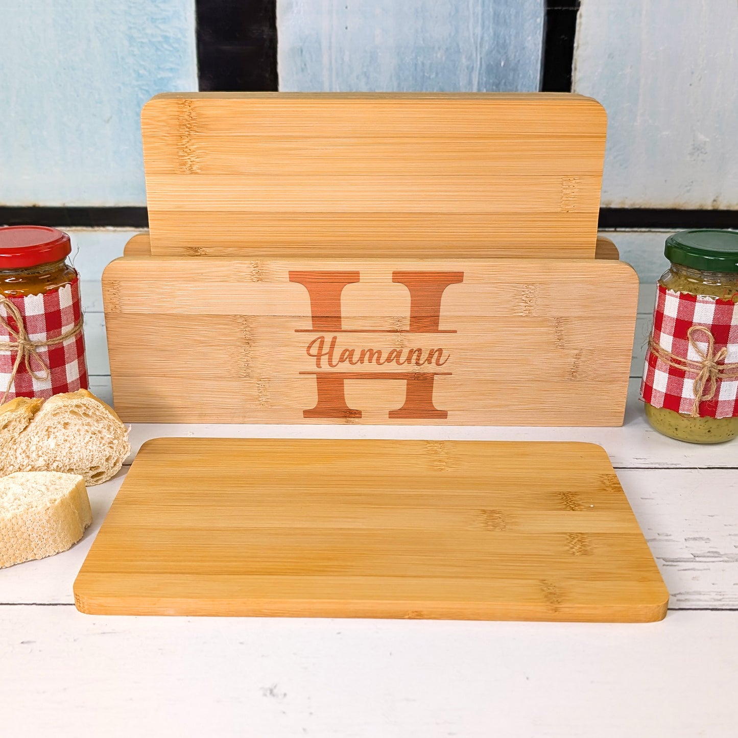 Brettchenständer mit fünf Bambusbrettchen, graviert mit dem Buchstaben „H“ und Namen „Hamann“, dazu Brot und Gläser mit Brotaufstrich.