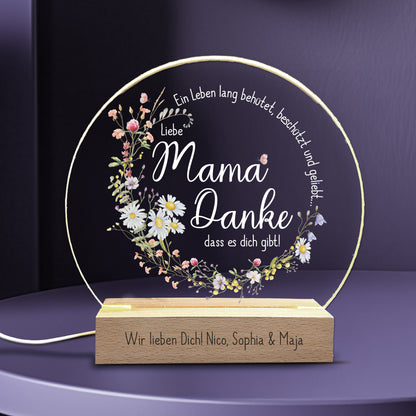 LED-Nachtlicht mit Blumenkranz und liebevollem Spruch für Mama: „Ein Leben lang behütet, beschützt und geliebt... Liebe Mama, Danke, dass es dich gibt!“. Die Acrylglasplatte wird von warmweißem LED-Licht beleuchtet und steht auf einem Holzsockel, der mit einer individuellen Gravur personalisiert werden kann. Ein besonderes Geschenk zum Muttertag oder als liebevolle Aufmerksamkeit.