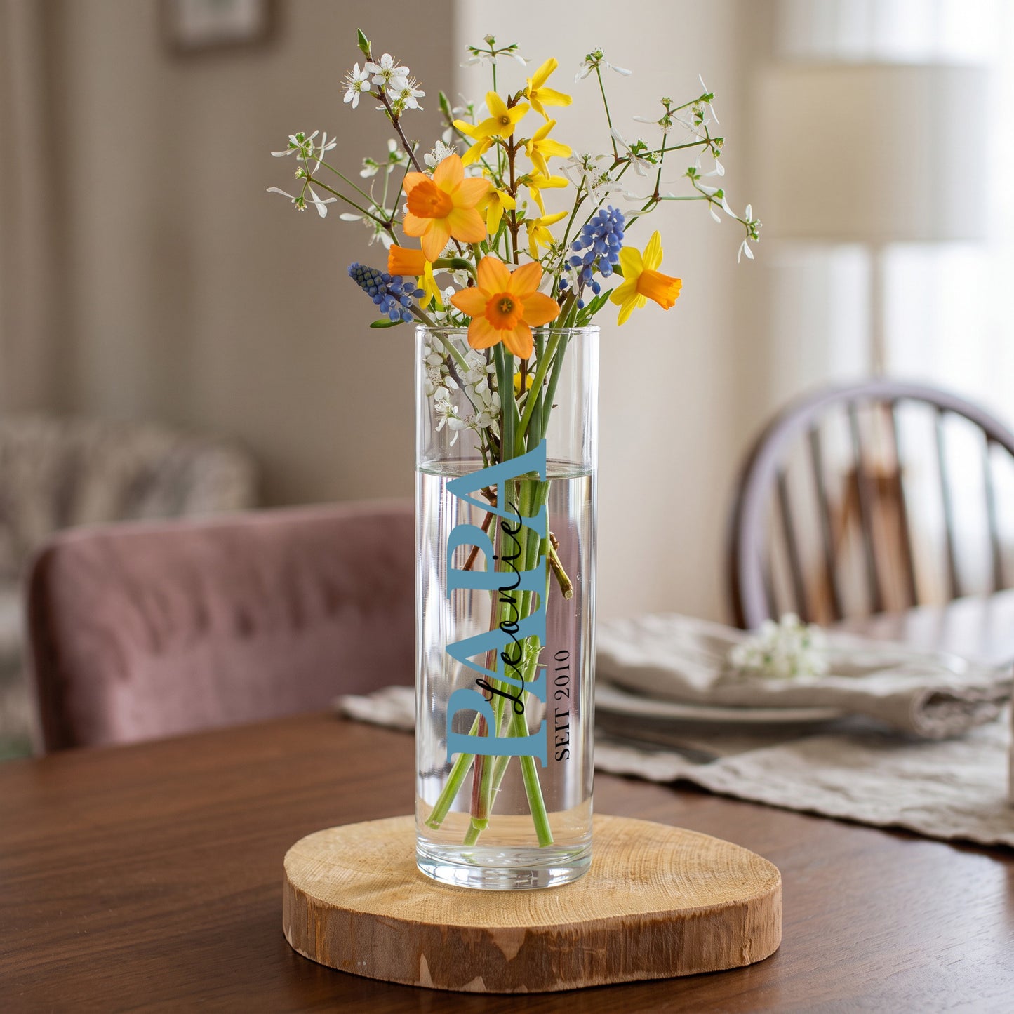 Personalisierte kleine Glasvase für Papa mit blauem Papa Schriftzug, Name Leonie und Jahreszahl 2010, dekoriert mit buntem Frühlingsblumenstrauß auf Holzscheibe