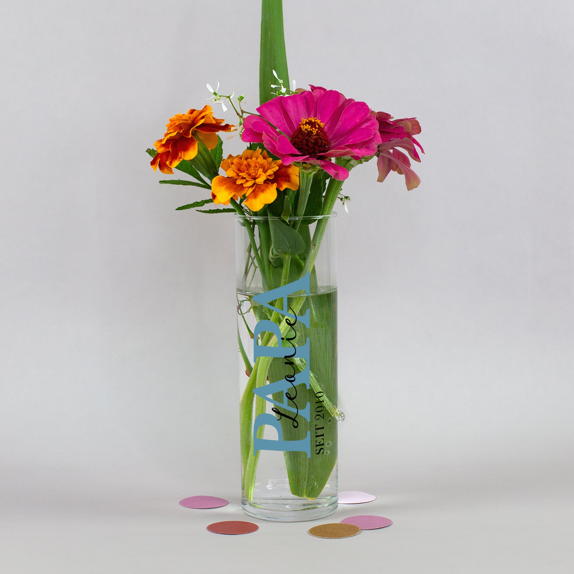 Kleine personalisierte Vase für Papa mit blauem Aufdruck, Wunschname und Jahreszahl, dekoriert mit farbenfrohem Blumenstrauß auf neutralem Hintergrund