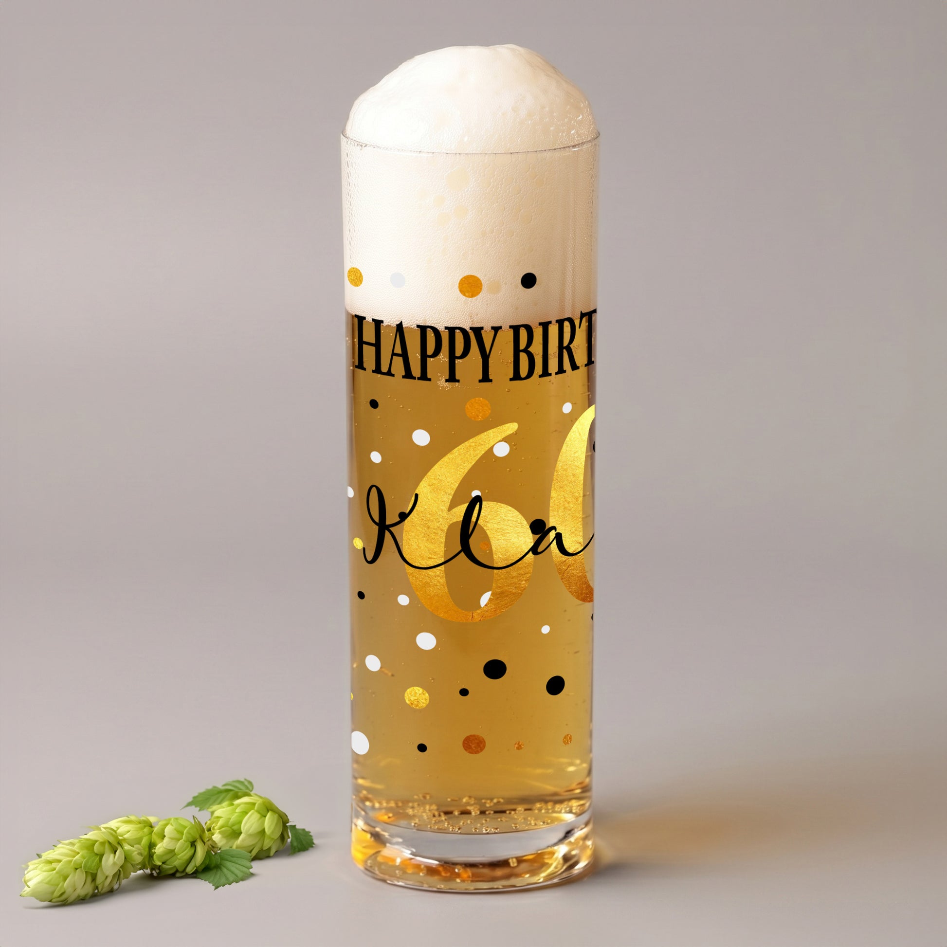 Kölschglas mit Geburtstagsmotiv, goldener Zahl 60 und personalisiertem Namen auf grauem Hintergrund, daneben grüne Hopfendolden.