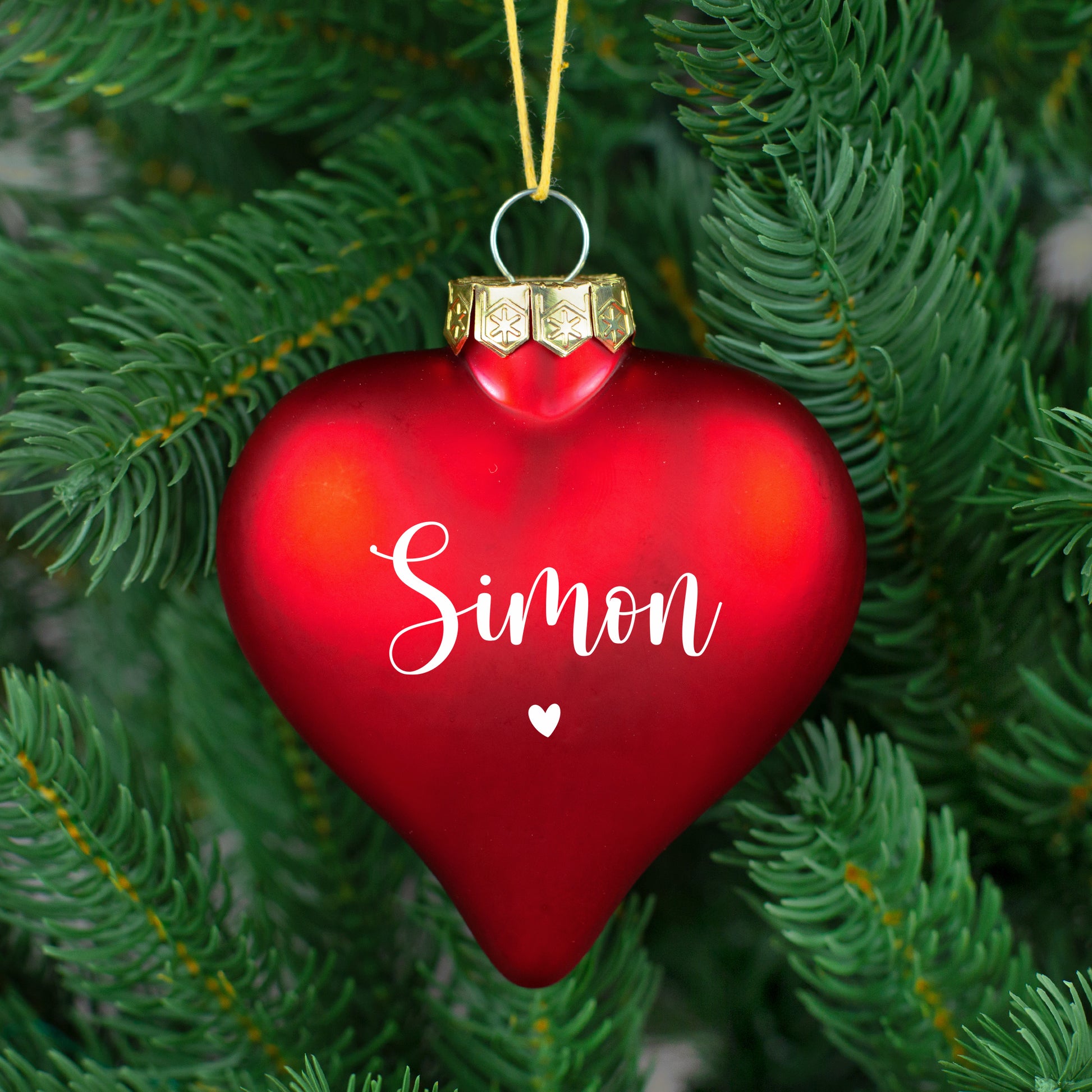 Rote, matte Herz-Christbaumkugel mit weißem Namen „Simon“ und kleinem Herz, an grünem Weihnachtsbaum.