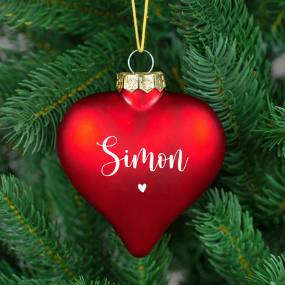 Glänzende rote Herz-Weihnachtskugel mit Name „Nicole“ auf weißem Fell, Tannenzweig im Anschnitt.