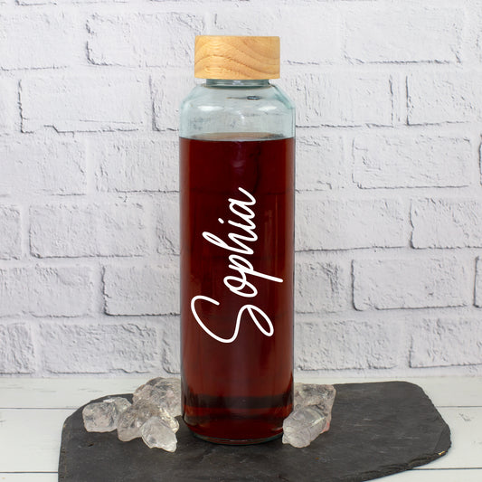 Personalisierte Trinkflasche aus recyceltem Glas mit weißem Namen Sophia, Holzdeckel und dunklem Getränk auf Schieferplatte