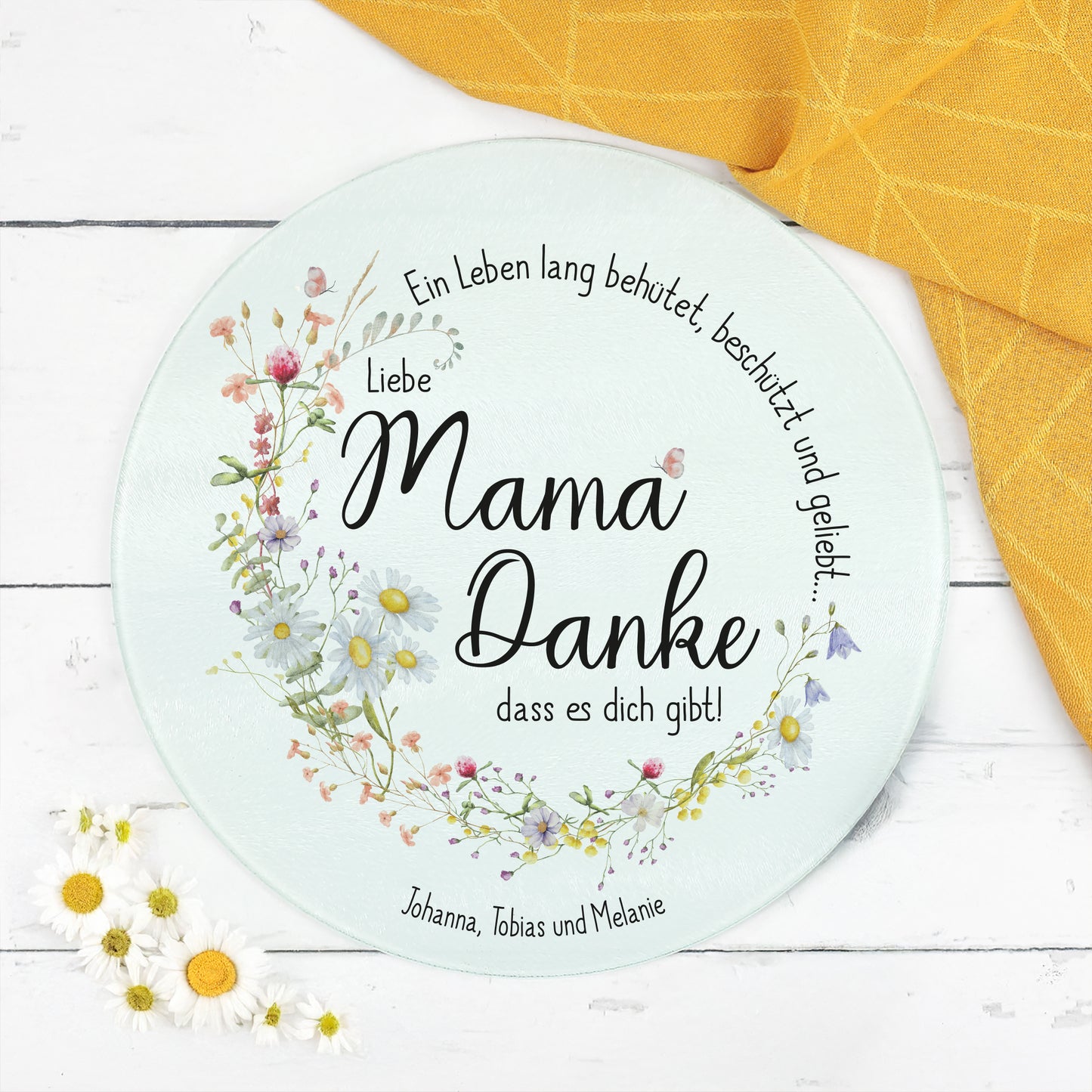 Rundes Glasbrettchen mit zartem Wiesenblumen-Kranz und dem Spruch „Ein Leben lang behütet, beschützt und geliebt…“. In der Mitte steht die Botschaft „Liebe Mama, Danke, dass es dich gibt!“. Am unteren Rand eine personalisierte Textzeile mit „Johanna, Tobias und Melanie“. Das Brett liegt auf einer weißen Holzoberfläche mit dekorativen Gänseblümchen und einem gelben Küchentuch.