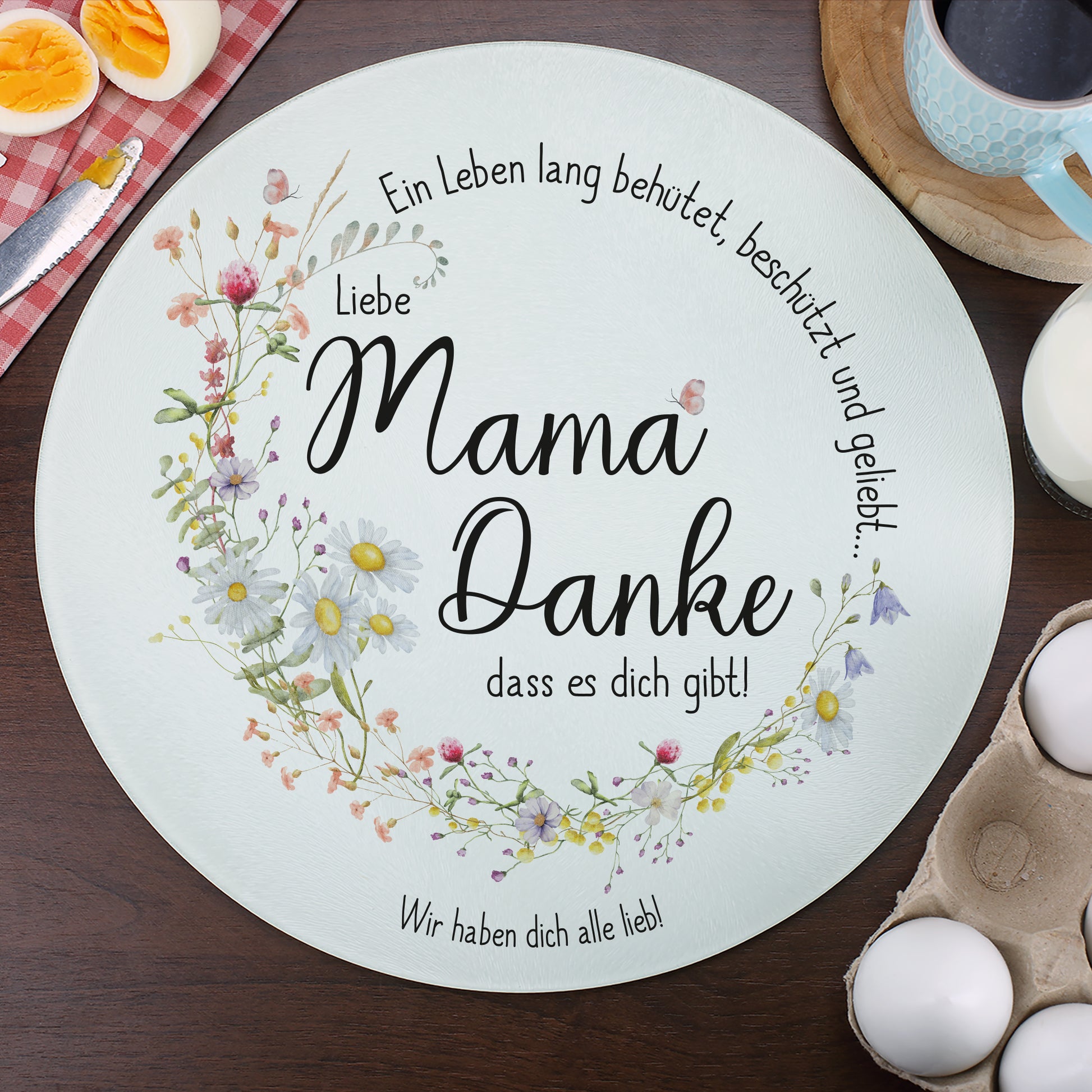 Rundes Glasbrettchen mit Wiesenblumen-Kranz und liebevollem Spruch für Mama, dekoriert auf einem Frühstückstisch mit Kaffee, Eiern und einem roten Küchentuch. Der personalisierte Wunschtext lautet „Wir haben dich alle lieb!“. Das Glasbrett ist vielseitig nutzbar als Servier- oder Schneidebrett.