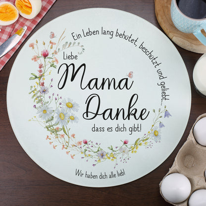 Rundes Glasbrettchen mit Wiesenblumen-Kranz und liebevollem Spruch für Mama, dekoriert auf einem Frühstückstisch mit Kaffee, Eiern und einem roten Küchentuch. Der personalisierte Wunschtext lautet „Wir haben dich alle lieb!“. Das Glasbrett ist vielseitig nutzbar als Servier- oder Schneidebrett.