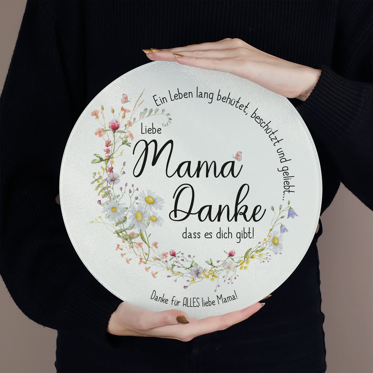 Eine Person hält das Glasbrettchen mit beiden Händen vor den Körper. Das transparente Material mit strukturierter Oberfläche ist gut sichtbar. Die Botschaft „Mama, Danke, dass es dich gibt!“ ist von einem detailreichen Blumenkranz und dem Schriftzug „Ein Leben lang behütet, beschützt und geliebt…“ umrahmt. Der Wunschtext unten lautet „Danke für ALLES liebe Mama!“.