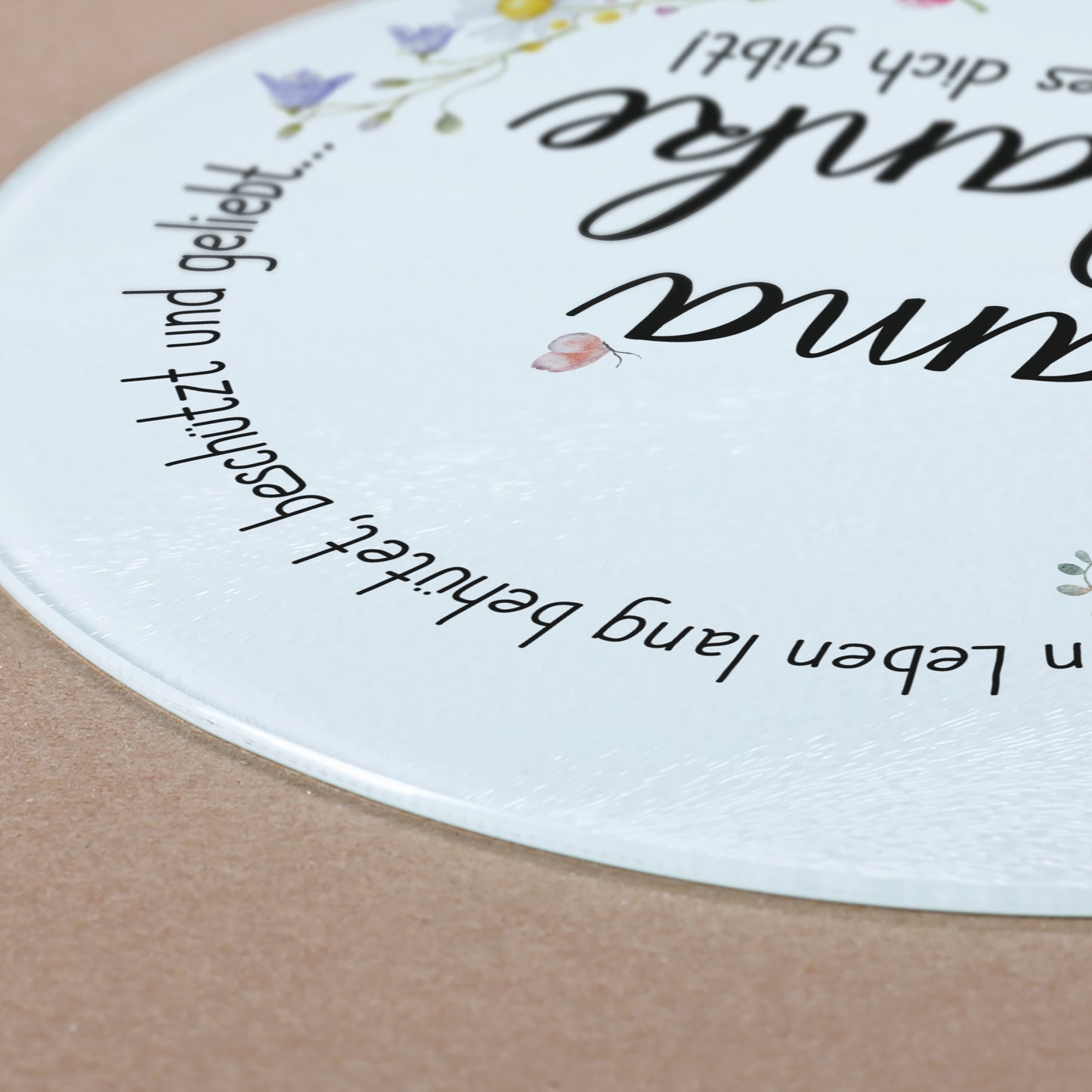 Nahaufnahme des Glasbrettchens, das auf einer hellbraunen Unterlage liegt. Die leicht erhabene Struktur der geriffelten Glasoberfläche ist erkennbar. Der Spruch „Ein Leben lang behütet, beschützt und geliebt…“ zieht sich sanft um den Rand des Bretts. Ein kleiner Schmetterling ergänzt das liebevolle Design.