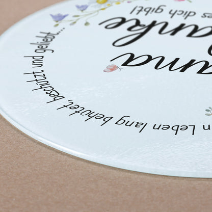 Nahaufnahme des Glasbrettchens, das auf einer hellbraunen Unterlage liegt. Die leicht erhabene Struktur der geriffelten Glasoberfläche ist erkennbar. Der Spruch „Ein Leben lang behütet, beschützt und geliebt…“ zieht sich sanft um den Rand des Bretts. Ein kleiner Schmetterling ergänzt das liebevolle Design.