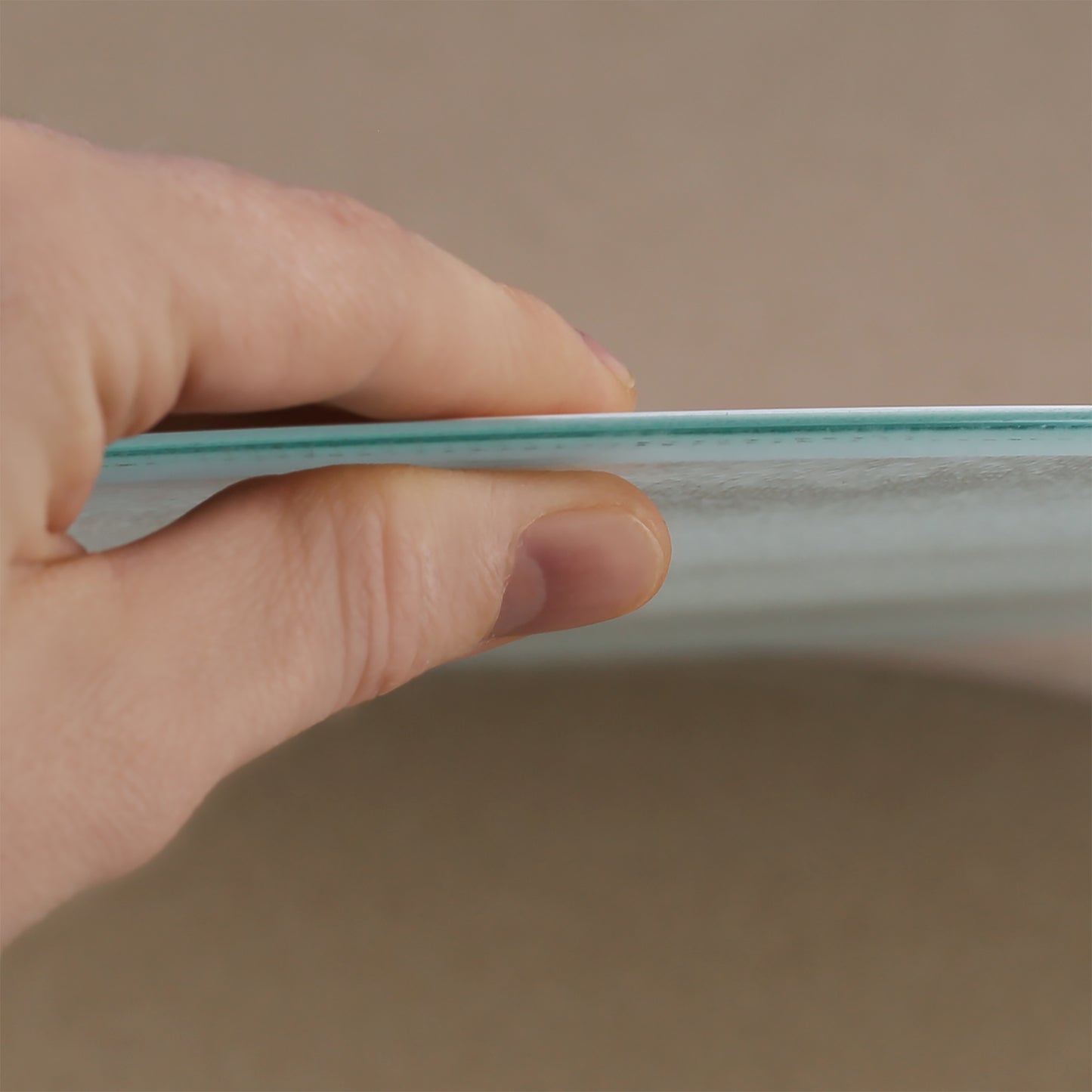Nahaufnahme einer Hand, die das Glasbrettchen an der Kante hält. Die dünne, stabile Glasplatte ist erkennbar, ebenso wie die leicht strukturierte Oberfläche. Das Bild zeigt die hochwertige Verarbeitung und das schlanke Design des Bretts.