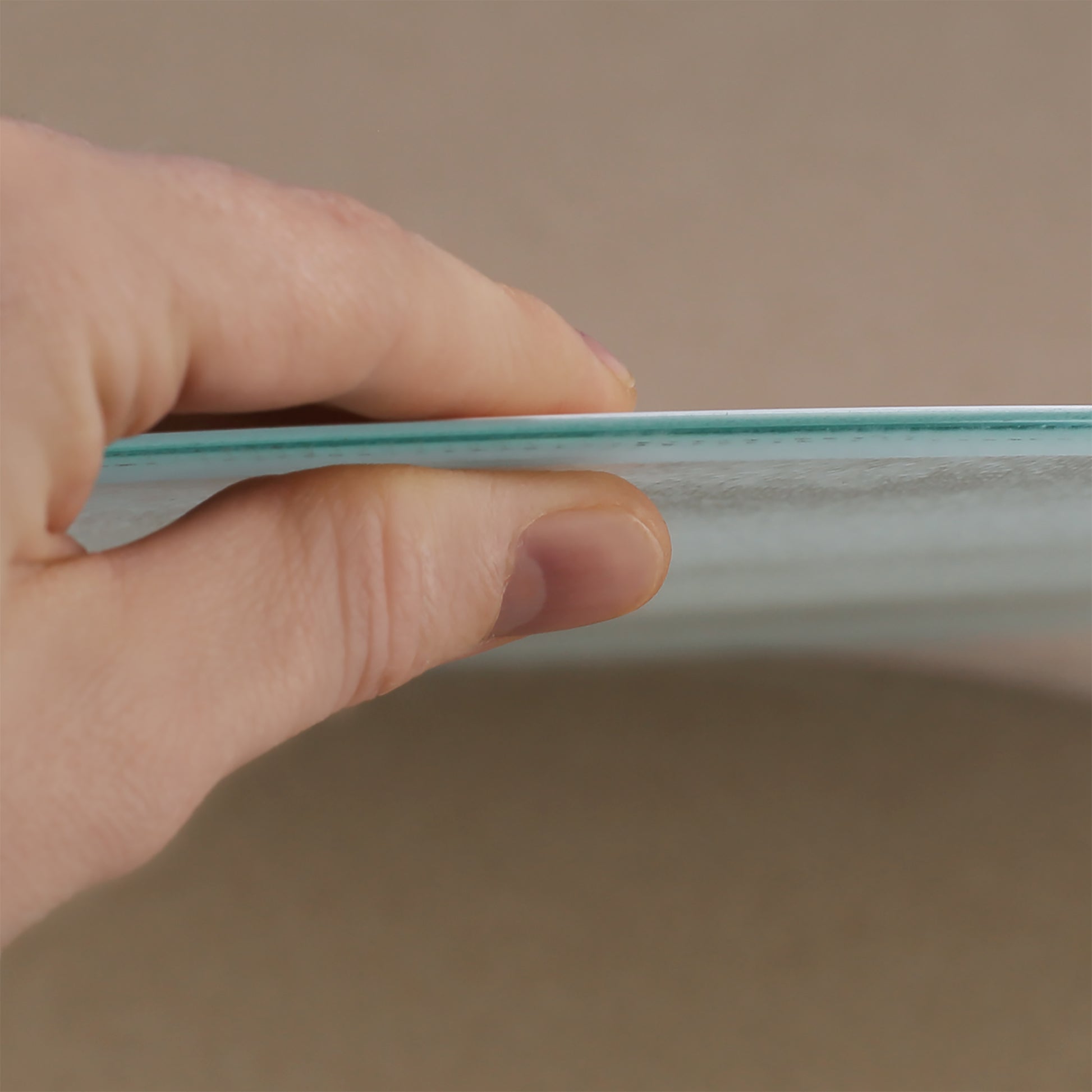 Nahaufnahme einer Hand, die das Glasbrettchen an der Kante hält. Die dünne, stabile Glasplatte ist erkennbar, ebenso wie die leicht strukturierte Oberfläche. Das Bild zeigt die hochwertige Verarbeitung und das schlanke Design des Bretts.